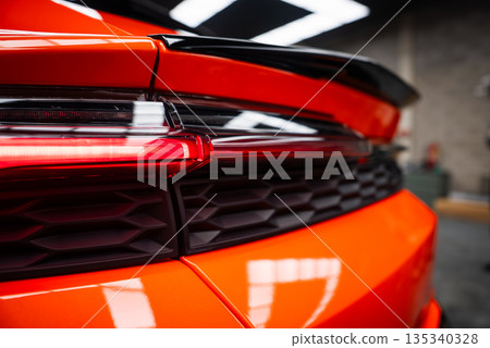 Close view of an orange Lamborghini Urus SE in a workshop, focusing on taillight, black honeycomb vent, and sculpted spoiler with overhead lights reflecting on glossy paint. Close view of an orange Lamborghini Urus SE in a workshop, focusing on taillight, black honeycomb vent, and sculpted spoiler with overhead lights reflecting on glossy paint. 135340328