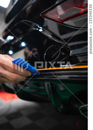A car technician uses a blue squeegee to align an orange vinyl stripe on a glossy black sports car rear bumper in a workshop, tail light and dual exhaust visible. A car technician uses a blue squeegee to align an orange vinyl stripe on a glossy black sports car rear bumper in a workshop, tail light and dual exhaust visible. 135340330