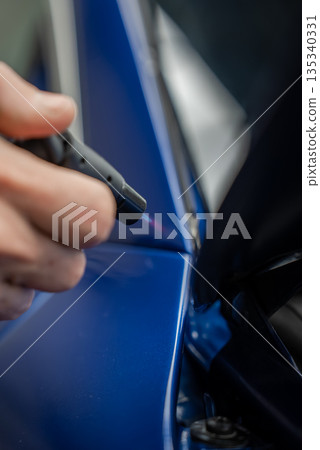 A technician moves a heat pen along a panel gap on a glossy blue luxury car in a dealership service bay. Soft lighting reveals precise detailing and a metallic finish. A technician moves a heat pen along a panel gap on a glossy blue luxury car in a dealership service bay. Soft lighting reveals precise detailing and a metallic finish. 135340331