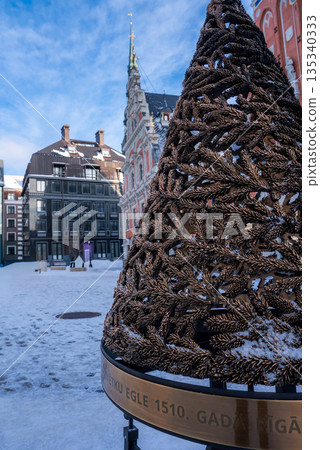 A bronze fir branch sculpture with snow and a ring marked 1510 stands in Riga Old Town, with House of the Blackheads and gabled facades under a crisp sky. A bronze fir branch sculpture with snow and a ring marked 1510 stands in Riga Old Town, with House of the Blackheads and gabled facades under a crisp sky. 135340333