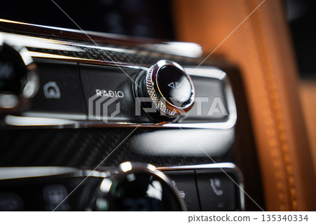 Close interior view shows a knurled chrome radio knob with arrow icon and RADIO label, carbon fiber, polished metal, and tan leather, in soft shallow depth lighting. 135340334