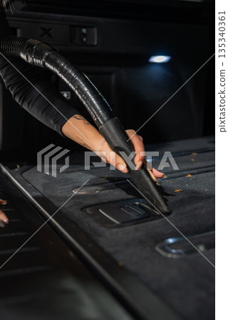A gloved person vacuums a carpeted cargo area, using a crevice tool around tie down latches and seat release handles, lit by an LED trunk light, showing debris removal. A gloved person vacuums a carpeted cargo area, using a crevice tool around tie down latches and seat release handles, lit by an LED trunk light, showing debris removal. 135340361