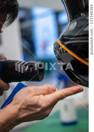 A car worker uses a heat gun to apply vinyl wrap on a glossy black panel with an orange stripe. Indoor workshop, green floor mat, blurred tools, shallow depth of field. 135340365