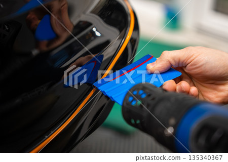 A car technician uses a blue squeegee and a heat gun to fit an orange pinstripe on a glossy black bumper edge in an indoor auto wrap workshop, emphasizing precision. 135340367