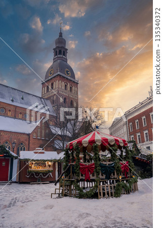 Snowy Christmas market in Riga, Latvia, shows carousel, wooden stalls, garlands, and the Riga Cathedral clock tower at dusk as locals and visitors gather. Snowy Christmas market in Riga, Latvia, shows carousel, wooden stalls, garlands, and the Riga Cathedral clock tower at dusk as locals and visitors gather. 135340372