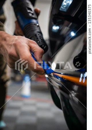 A car technician uses a blue squeegee and a heat gun on a glossy black vehicle indoors. Hands focus on an orange strip, with checkered floor and bright task lights visible. A car technician uses a blue squeegee and a heat gun on a glossy black vehicle indoors. Hands focus on an orange strip, with checkered floor and bright task lights visible. 135340375