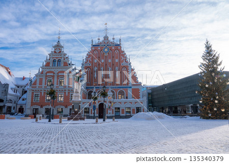 Riga, Latvia, Town Hall Square shows the House of the Blackheads, the guild, and the Roland statue. Snow covers the square while a lit Christmas tree glows on the right. 135340379