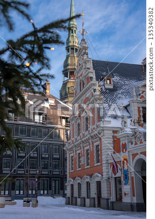 House of the Blackheads stands in Riga, Latvia as snow dusts cobblestones. St. Peter's Church spire rises behind. A Latvian flag and fairy lights signal Christmas afternoon. 135340382