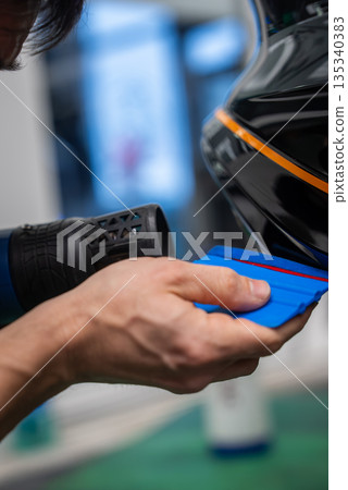 Close up shows a worker applying vinyl wrap to a glossy black panel using a heat gun and blue squeegee, forming around an edge with an orange stripe in a workshop. 135340383