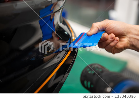 A worker smooths vinyl wrap on a glossy black bumper with a blue squeegee. Orange alignment tape marks the gap, and a heat gun lies nearby. Bright, shallow depth. 135340386