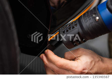 Close up shows a car technician aligning an orange strip and heating a vinyl wrap around a wheel arch in a workshop, with focused lighting revealing precise reflections. Close up shows a car technician aligning an orange strip and heating a vinyl wrap around a wheel arch in a workshop, with focused lighting revealing precise reflections. 135340387