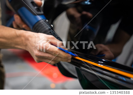 Close up of hands in an auto service bay applying an orange vinyl stripe on a glossy black panel, using a heat gun and blue squeegee under bright diffused lighting. Close up of hands in an auto service bay applying an orange vinyl stripe on a glossy black panel, using a heat gun and blue squeegee under bright diffused lighting. 135340388