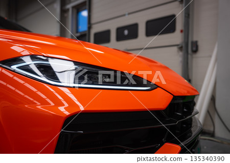 Close up shows the front left headlight, LED daytime running light, and grille of an orange Lamborghini Urus SE inside a workshop with tools and garage doors. Close up shows the front left headlight, LED daytime running light, and grille of an orange Lamborghini Urus SE inside a workshop with tools and garage doors. 135340390