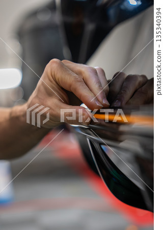 Close view shows a car technician aligning an orange trim tool on a glossy black panel in an auto workshop, indicating film or vinyl work under soft indoor lighting. Close view shows a car technician aligning an orange trim tool on a glossy black panel in an auto workshop, indicating film or vinyl work under soft indoor lighting. 135340394