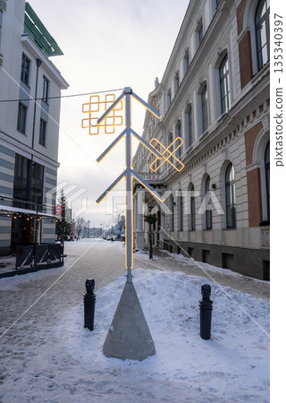 A neon tree sculpture with Baltic motifs stands on a snowy cobblestone street in Riga, Latvia, framed by 19th century facades, in soft winter daylight. A neon tree sculpture with Baltic motifs stands on a snowy cobblestone street in Riga, Latvia, framed by 19th century facades, in soft winter daylight. 135340397