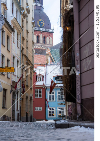 A narrow snow dusted street in Riga Old Town leads to Riga Cathedral clock tower and copper dome, with cream, red, pastel blue facades and Latvian flags in winter. A narrow snow dusted street in Riga Old Town leads to Riga Cathedral clock tower and copper dome, with cream, red, pastel blue facades and Latvian flags in winter. 135340399