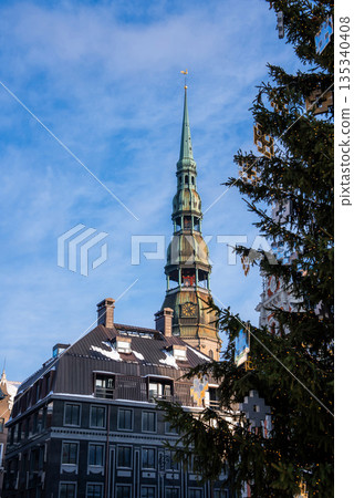 St. Peter's Church tower rises above Riga Old Town, Latvia. A Christmas tree glows with lights and ornaments, snow rests on dark roofs, and a clock and weather vane show in daylight. 135340408