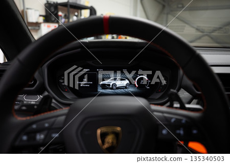 Close up from the driver seat shows the Lamborghini crest wheel and red marker, with the Urus digital cluster active, in a workshop under soft indoor lighting. 135340503