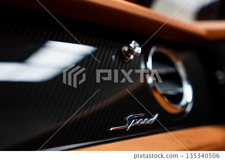 Close interior view shows carbon fiber dashboard trim, Speed script badge, and chrome air vent. Warm tan leather and soft showroom lighting emphasize premium detail. 135340506