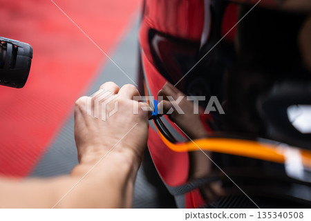 A worker uses a thin blue tool along a glossy black car panel edge in an indoor service bay with red and gray flooring, bright orange stripe reflections, and soft, shallow focus. 135340508