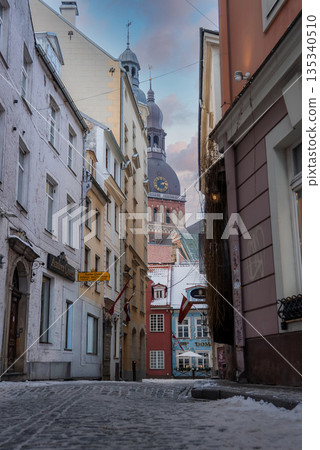 A narrow cobblestone lane in Riga Old Town leads to Riga Cathedral. Snow rests on edges and sills. Latvian flags and signs appear. Soft cool daylight sets a calm mood. A narrow cobblestone lane in Riga Old Town leads to Riga Cathedral. Snow rests on edges and sills. Latvian flags and signs appear. Soft cool daylight sets a calm mood. 135340510
