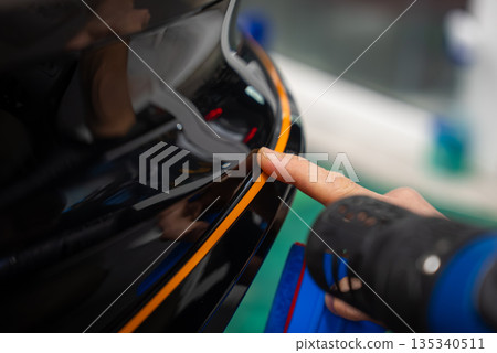 A car worker aligns an orange vinyl stripe on a glossy black bumper, using a heat gun to conform the film. Bright task lighting and tools surround the tight, shallow view. A car worker aligns an orange vinyl stripe on a glossy black bumper, using a heat gun to conform the film. Bright task lighting and tools surround the tight, shallow view. 135340511