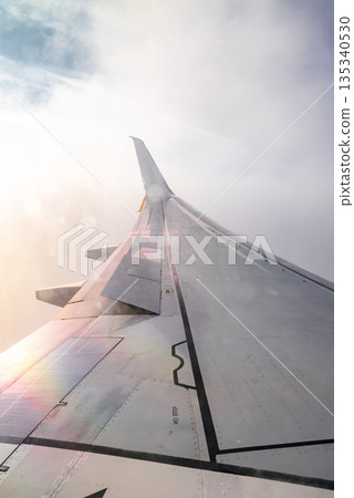 Aerial view from a passenger window shows an airplane wing with extended flaps cutting through clouds near Liverpool. Sunlight diffuses across metal during daytime. 135340530