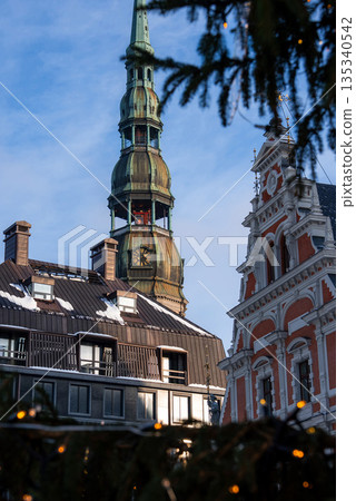 Street level view shows St. Peter's Church spire and clock above facades in Riga, Latvia. House of the Blackheads appears right, with snow, evergreens, and fairy lights. Street level view shows St. Peter's Church spire and clock above facades in Riga, Latvia. House of the Blackheads appears right, with snow, evergreens, and fairy lights. 135340542