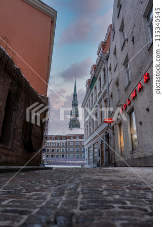 Narrow cobblestone alley in Riga Old Town opens to a square, pastel facades, neon VOLMAR sign, and St. Peter's Church spire in soft winter daylight with light snow. 135340545