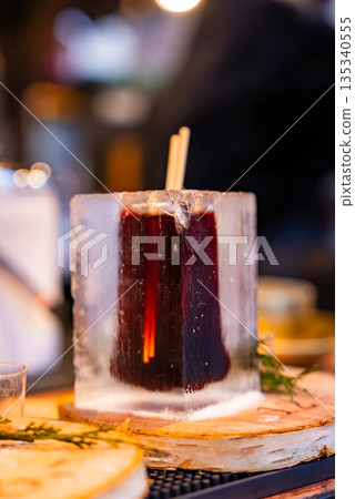 A steaming mulled wine in a clear ice block sits on a birch coaster in Riga, Latvia. Warm market lights blur behind, with sprigs of greenery suggesting festive winter mood. A steaming mulled wine in a clear ice block sits on a birch coaster in Riga, Latvia. Warm market lights blur behind, with sprigs of greenery suggesting festive winter mood. 135340555