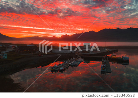 A small Icelandic harbor during a red and orange sunset, with docked boats, calm reflective water, rugged mountain silhouettes, and nearby buildings. A small Icelandic harbor during a red and orange sunset, with docked boats, calm reflective water, rugged mountain silhouettes, and nearby buildings. 135340558