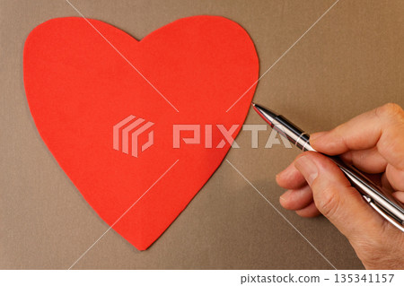 Man hand writing on blank red heart card with pen on brown background. Man hand writing on blank red heart card with pen on brown background. 135341157