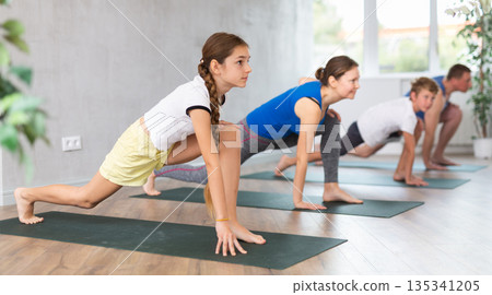 Pleasant preteen girl practicing variation of warrior pose of yoga with her family 135341205