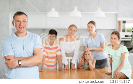 Smiling dad poses in cozy kitchen, large family with four children and wife are standing behind Smiling dad poses in cozy kitchen, large family with four children and wife are standing behind 135341337