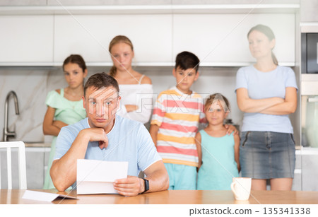 Upset father reads and looks through financial documents. Sad wife with children in background 135341338