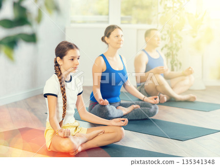 School children and their parents sitting on mats in Lotus pose during family yoga training in gym fitness center indoors 135341348