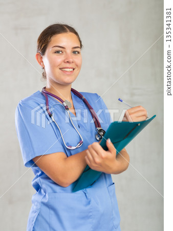 Positive young woman general practitioner in blue uniform filling out clipboard with medical records. Studio photoshoot on gray background 135341381