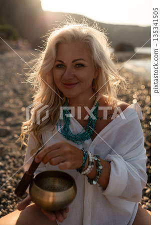 Woman, Meditation, Beach: Blonde woman playing singing bowl on pebble beach at tranquil sunset, promoting wellness and spiritual practice Woman, Meditation, Beach: Blonde woman playing singing bowl on pebble beach at tranquil sunset, promoting wellness and spiritual practice 135341505