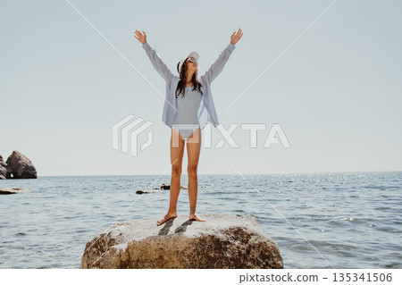 Woman Beach Joy, young woman raises arms on rock in sparkling sea under clear sky, feeling free. 135341506