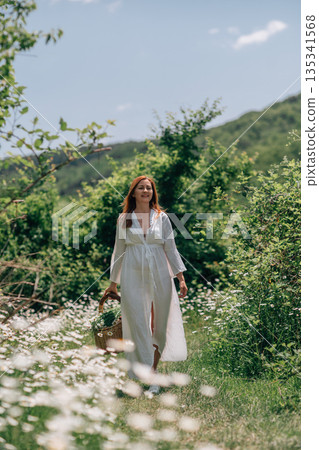 Woman Basket Daisies: Summer day; red-haired woman carries flower basket in chamomile field for foraging. Woman Basket Daisies: Summer day; red-haired woman carries flower basket in chamomile field for foraging. 135341568
