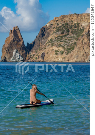 Paddleboard woman sea, a person kneeling on a stand-up paddleboard in the clear ocean with majestic rocky cliffs. Paddleboard woman sea, a person kneeling on a stand-up paddleboard in the clear ocean with majestic rocky cliffs. 135341647
