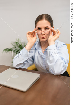 Businesswoman glasses professional looking seriously at camera while adjusting eyewear at a desk Businesswoman glasses professional looking seriously at camera while adjusting eyewear at a desk 135341648