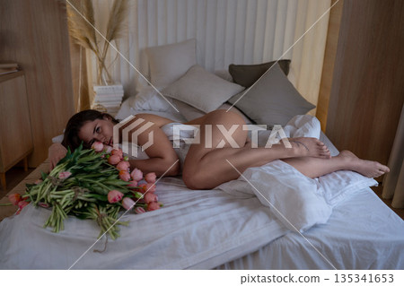 Woman, tulips, bedroom. Young brunette woman relaxing on white sheets with bouquet of pink flowers Woman, tulips, bedroom. Young brunette woman relaxing on white sheets with bouquet of pink flowers 135341653