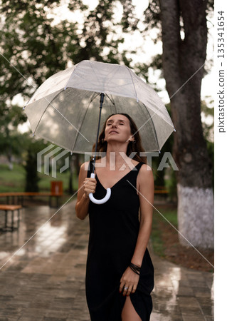 Woman, rain, umbrella. Serene woman enjoying refreshing drizzle with a clear umbrella on a wet park path. Woman, rain, umbrella. Serene woman enjoying refreshing drizzle with a clear umbrella on a wet park path. 135341654