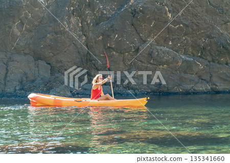 Kayak woman paddling, enjoying summer adventure in clear coastal water near rocky cliff Kayak woman paddling, enjoying summer adventure in clear coastal water near rocky cliff 135341660