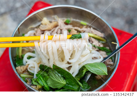 Close-up of chopsticks holding flat rice noodles (Banh Pho) from a metal bowl of Vietnamese noodle soup  with meat, herbs, and basil on a red plastic stool. 135342437