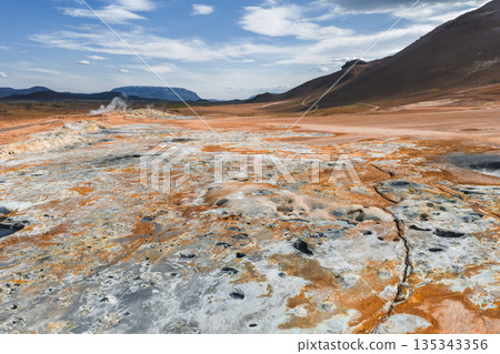 The geothermal area of Hverir near Lake Myvatn in Iceland features orange and white mineral streaked ground, steam vents, fumaroles, and rolling hills. 135343356