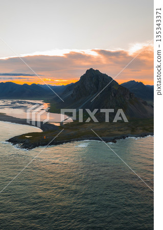 Aerial view of Vestrahorn mountain on Iceland's Stokksnes Peninsula, surrounded by black sand beaches, reflective waters, and a vivid sunset sky. Aerial view of Vestrahorn mountain on Iceland's Stokksnes Peninsula, surrounded by black sand beaches, reflective waters, and a vivid sunset sky. 135343371