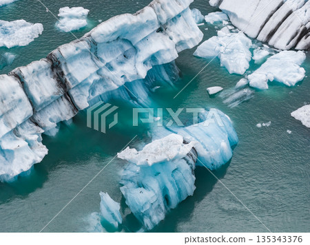 Large icebergs with blue and white hues, some streaked with volcanic ash, float in a vivid turquoise glacial lagoon, showcasing Iceland's geology. 135343376