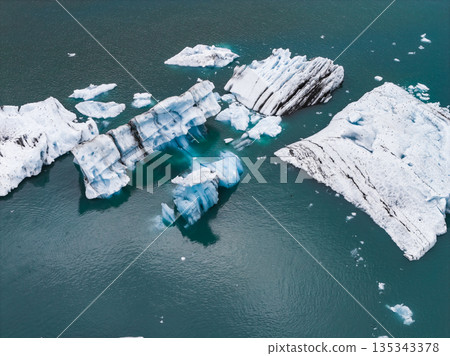 Large icebergs with white, blue, and dark streaks float in a calm, teal glacial lagoon. The water reflects the icy formations, creating a serene scene. Large icebergs with white, blue, and dark streaks float in a calm, teal glacial lagoon. The water reflects the icy formations, creating a serene scene. 135343378